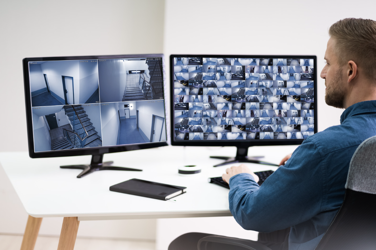 Man sitting at desk looking at computer screens with multiple CCTV footage.